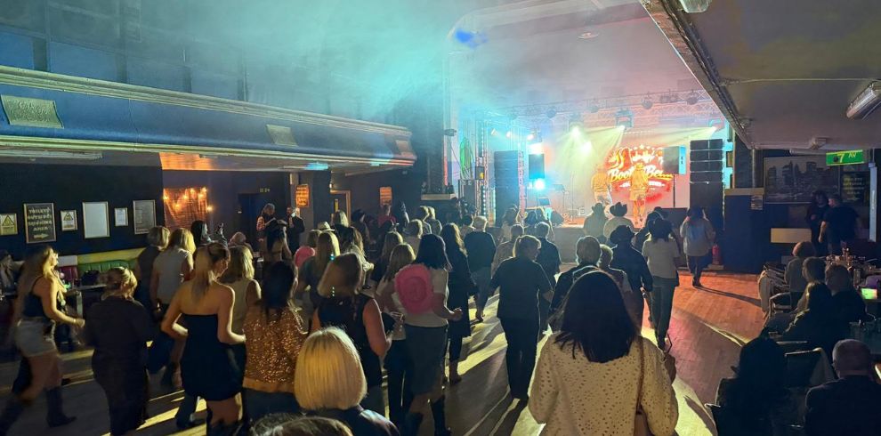 Wide view of a busy indoor venue with a crowd facing a stage, colourful lights and smoke effects, and a live performer at the front.