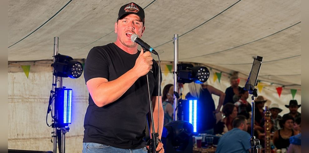 Performer in a black t-shirt and cap singing into a microphone on a small stage, with lighting equipment and seated audience visible behind.