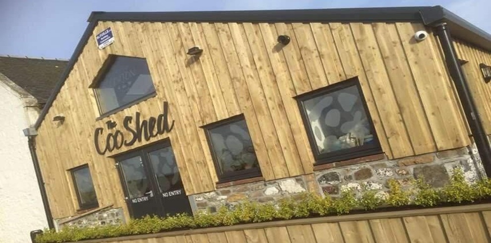 Exterior of The Coo Shed café, a modern wooden-clad building with stone base detailing, featuring angular windows and a central entrance with “Coo Shed” signage above the doors, set against a clear sky with a fenced foreground.