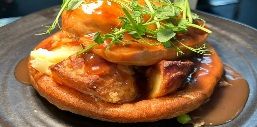 A close-up of a plated dish featuring a golden Yorkshire pudding filled with pieces of roasted meat and creamy mashed potatoes, topped with a portion of glazed meat covered in rich brown gravy. The gravy is generously poured over the entire dish and pools on a dark-colored plate. Fresh green pea shoots are scattered on top as garnish, adding contrast to the warm brown and golden tones of the food. The focus is tight on the dish, with the background blurred.