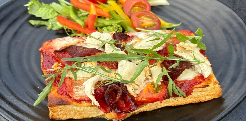 A close-up of a rectangular slice of pizza on a dark plate, topped with melted cheese, tomato sauce, pieces of crispy bacon, and chunks of cooked chicken. Fresh rocket leaves are scattered over the top. To the side sits a small mixed salad with green lettuce, sliced red and yellow peppers, and halved cherry tomatoes. The background is blurred, keeping the focus on the plated food.