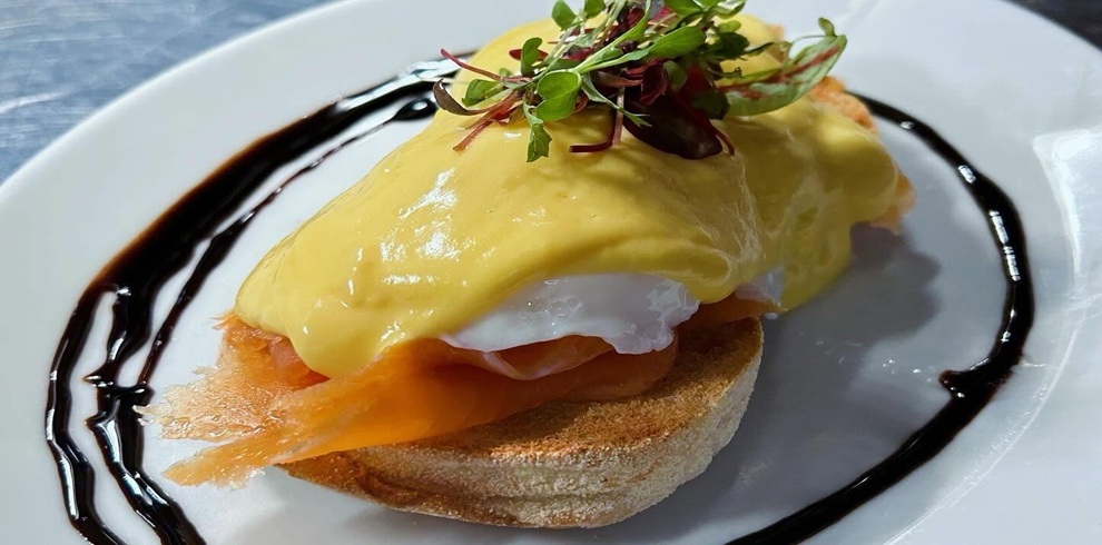A close-up of a plated eggs Benedict dish featuring a toasted English muffin topped with folded slices of smoked salmon, a poached egg, and a generous layer of smooth, yellow hollandaise sauce. The dish is garnished with a small cluster of fresh microgreens on top. A dark balsamic-style glaze is drizzled in a decorative ring around the edge of the white plate, contrasting with the light tones of the food. The background is softly blurred, keeping the focus on the dish’s textures and colors.