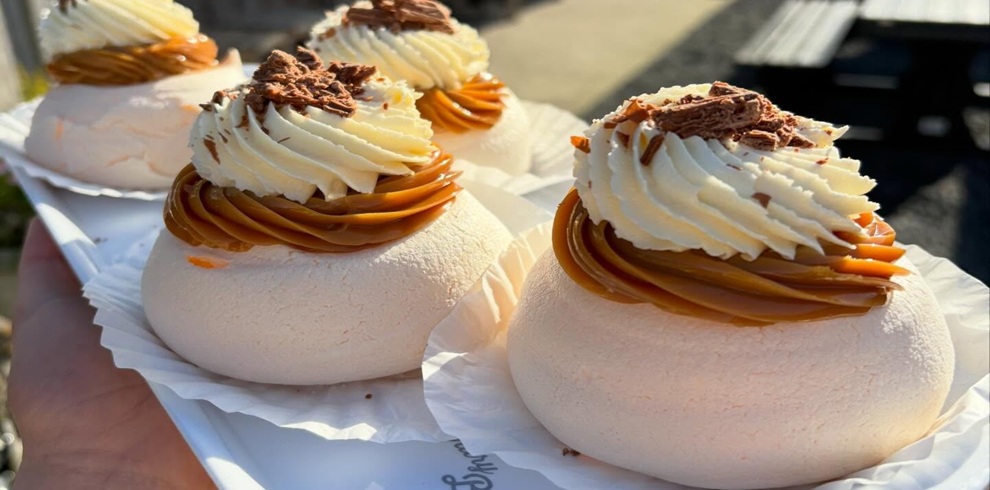 A close-up of several round meringue desserts arranged on a white tray, each topped with a swirl of caramel-colored sauce and a generous layer of piped whipped cream. Small chocolate flakes are sprinkled on top of the cream. The meringues are light beige with a slightly crisp exterior, each resting in a white paper case. A hand is visible holding the tray, and the background is softly blurred with outdoor seating faintly visible in sunlight.