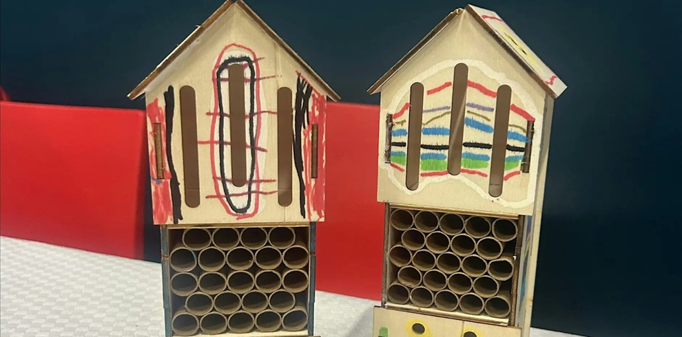 Two small, handmade wooden insect houses are placed side by side on a surface. Each structure has a pitched roof and a front section filled with neatly arranged hollow tubes, designed to provide shelter for insects. The upper portions are decorated with colourful, childlike drawings—one featuring bold black and red markings, and the other displaying horizontal stripes in multiple colours. The background includes a red and blue panel, creating a simple, bright setting that contrasts with the natural wood tones of the insect houses.