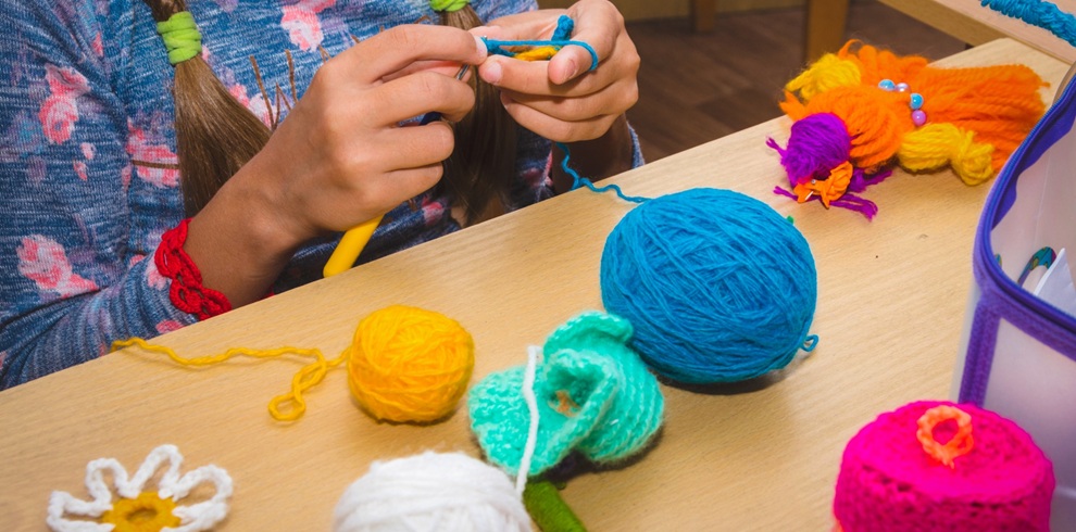 Close-up of a person’s hands crocheting with a yellow hook and bright blue yarn at a table. Several balls of yarn in different colors, including yellow, blue, and white, are scattered across the surface, along with small crocheted items such as flowers and shapes. A colorful crocheted bird-like figure and a container holding craft supplies sit nearby, suggesting a creative crochet or craft activity setting.