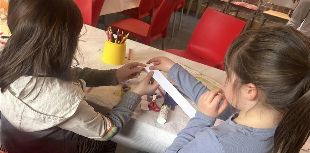 Two children sit at a table indoors, working together on a craft activity. One child holds a long strip of white paper while the other helps position it. On the table are coloured pencils stored in a yellow cup, a glue stick, scissors, and small craft materials scattered across the surface. Red chairs and additional tables are visible in the background, suggesting a classroom or workshop setting.
