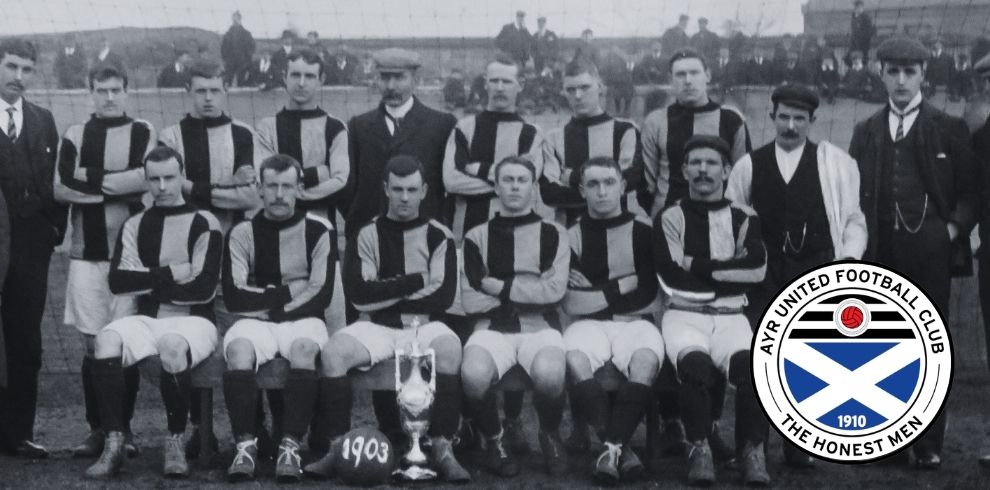 Black-and-white historic team photograph of an early Ayr United football squad posed in rows on a pitch. The players wear striped jerseys and shorts, with some standing behind and others seated in front. A football trophy is placed at the centre in the foreground, beside a ball marked “1903.” A crowd of spectators lines the background behind a low boundary wall. In the bottom right corner, a modern Ayr United Football Club crest featuring a blue and white saltire and the text “Ayr United Football Club – The Honest Men – 1910” is overlaid on the image.