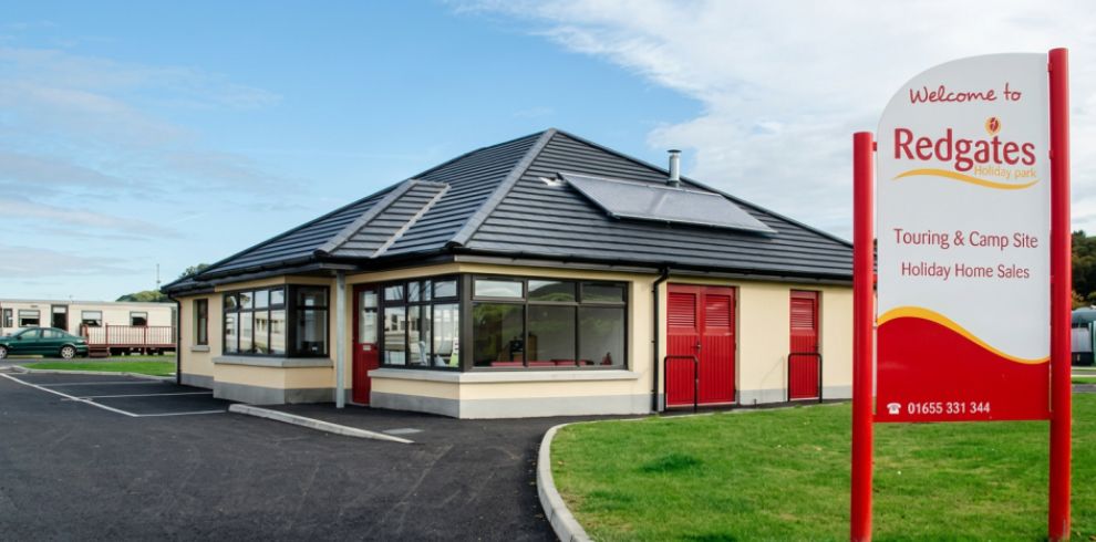 A modern reception building at Redgates Holiday Park, with cream walls, a grey tiled roof fitted with solar panels, and bright red doors and shutters. The building sits beside a paved driveway and parking area, with holiday caravans visible in the background. In the foreground, a large white and red sign reads “Welcome to Redgates Holiday Park – Touring & Camp Site, Holiday Home Sales” along with a phone number (01655 331 344). The scene is set under a clear blue sky with neatly maintained green grass surrounding the entrance.