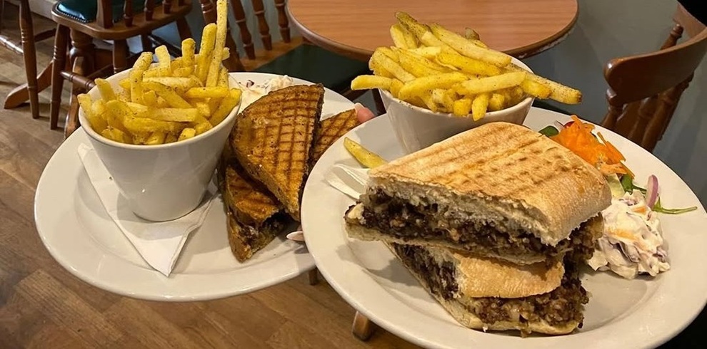 Two plates of café food on a wooden table: each plate holds a toasted panini sandwich, with crisp fries served in white cups. One sandwich is cut into triangles with visible grill marks, while the other is stacked high with a dark filling, served alongside coleslaw and a small salad garnish. Wooden chairs and another table are visible in the background, suggesting a casual dining setting.