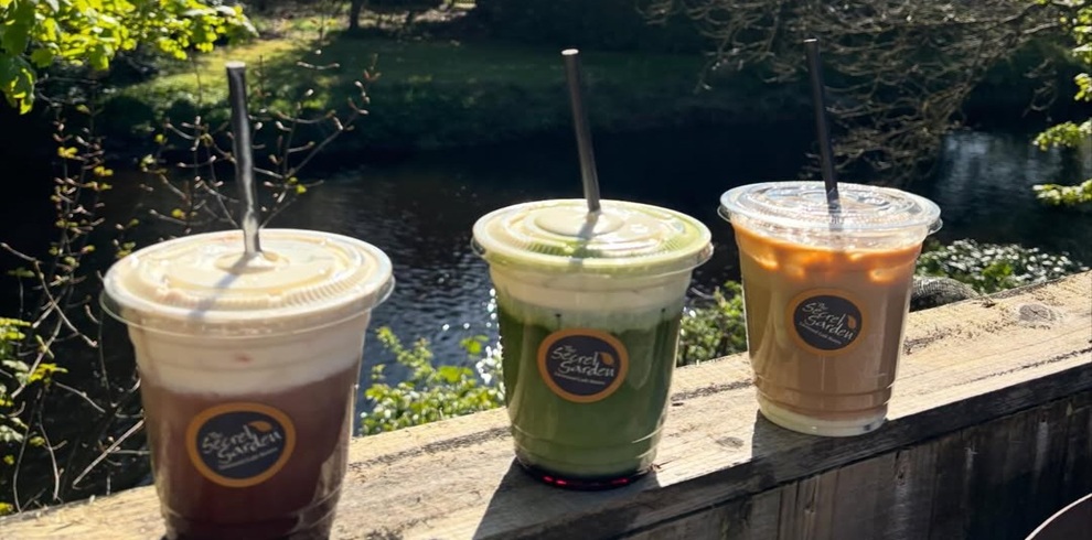 Three takeaway iced drinks sit on a wooden railing overlooking a calm river surrounded by greenery. Each clear plastic cup has a lid, a black straw, and a circular “Secret Garden” label. The drinks include a dark iced coffee with a creamy layer on top, a bright green layered beverage, and a light brown iced coffee or latte. Sunlight reflects off the water in the background, creating a bright, natural outdoor setting.