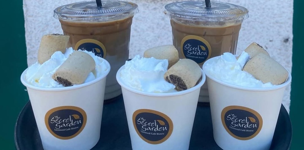 White takeaway cups filled with whipped cream and topped with wafer rolls sit on a black tray, placed in front of two clear plastic iced coffee cups with lids and straws. Each cup displays a circular logo reading “The Secret Garden Licensed Café Bistro.” The drinks are arranged outdoors against a light-colored wall, with soft natural lighting highlighting the creamy textures and light brown coffee.