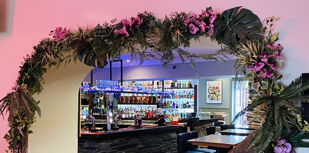 Interior view of Smith’s Bar & Restaurant in Ayr, featuring a decorated archway with greenery and purple flowers framing a modern bar area with illuminated shelves of spirits, beer taps, and wooden seating in a softly lit dining space.