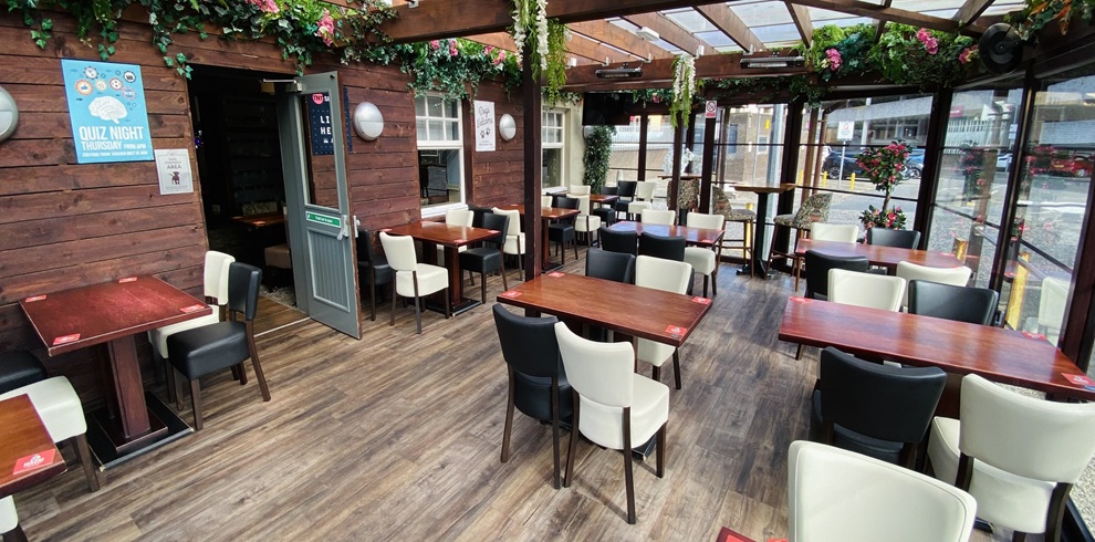 Indoor covered seating area at Smith’s Bar & Restaurant in Ayr, featuring wooden tables with black and cream chairs, wood-panelled walls, and a glass-roofed space decorated with hanging greenery and flowers, with natural light coming through large windows.