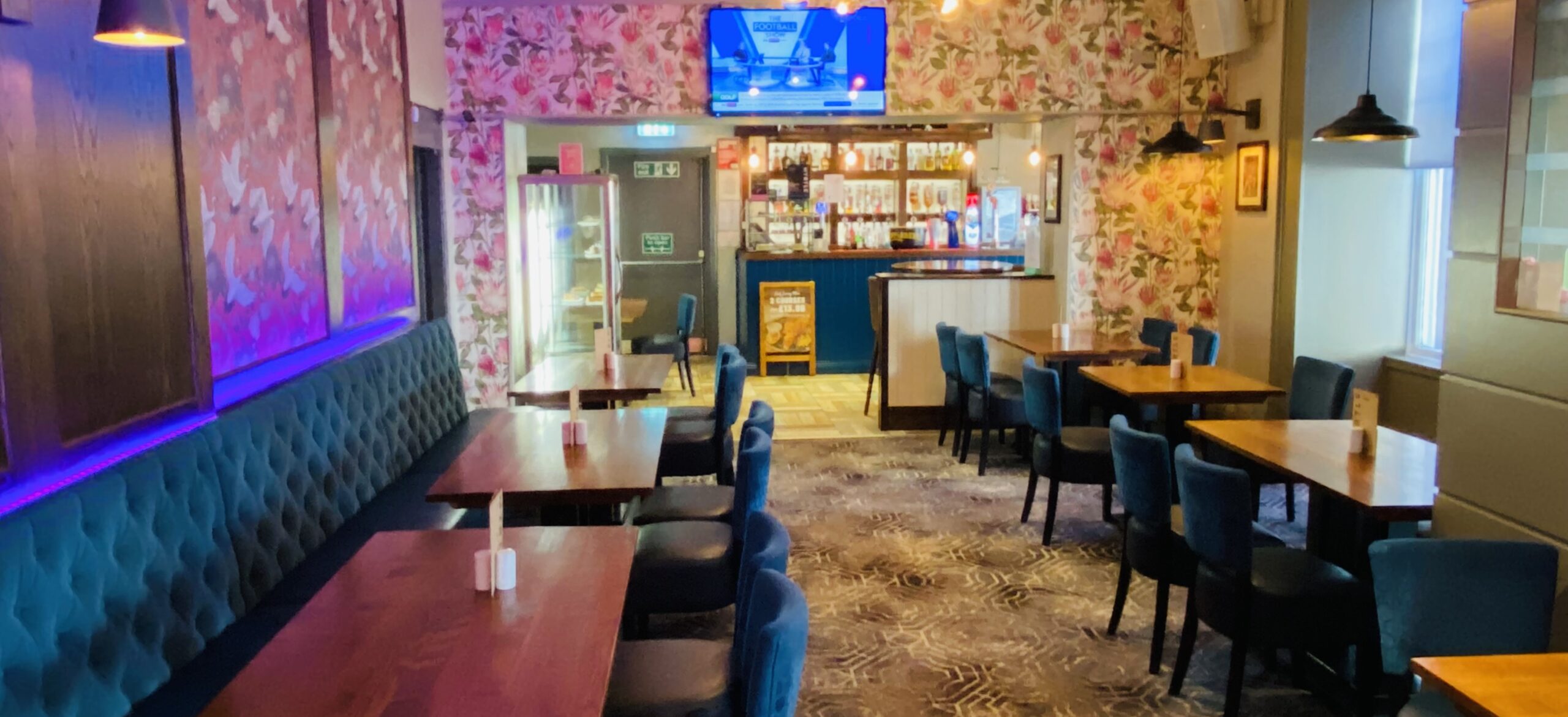 Interior dining area at Smith’s Bar & Restaurant in Ayr, featuring wooden tables, upholstered bench seating and chairs, floral wallpaper, and a view towards the bar with illuminated shelves, a drinks fridge, and a TV screen showing live sports above the counter.