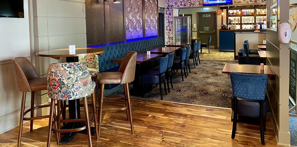Interior dining area at Smith’s Bar & Restaurant in Ayr, featuring wooden tables, upholstered bench seating and chairs, floral wallpaper, and a view towards the bar with illuminated shelves, a drinks fridge, and a TV screen showing live sports above the counter.