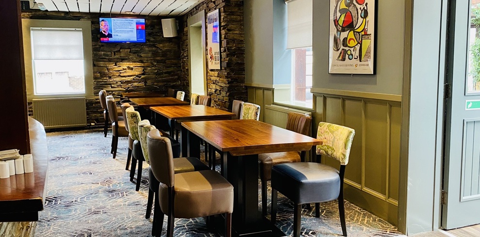 A cozy, modern pub interior with a row of rectangular wooden tables and cushioned chairs arranged along the right wall. The wall features decorative paneling painted in a muted green tone, with framed artwork and large windows letting in natural light. On the left side, a stone accent wall runs the length of the room, with a wall-mounted television displaying a news program. The floor has a patterned carpet in blue and gray tones, and the overall lighting is warm, creating an inviting dining atmosphere.