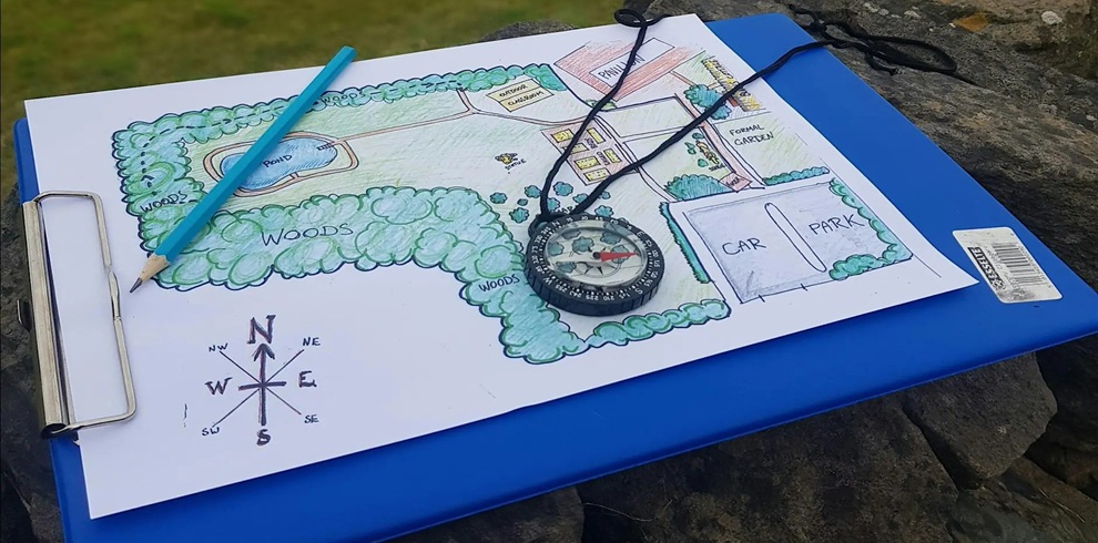 A clipboard rests on a rough stone surface outdoors, holding a hand-drawn map colored with greens and blues. The map shows labeled areas including “woods,” a small “pond,” roads, houses, and spaces marked “car park” and “formal garden.” A blue pencil lies diagonally across the paper, and a black compass with a cord is placed near the center of the map. In the lower-left corner of the page, a compass rose indicates directions (N, E, S, W). The background features blurred greenery, suggesting a natural setting.