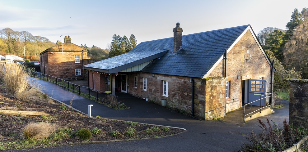 Stone and brick building with a dark slate roof, viewed from an elevated angle. A paved pathway and accessibility ramp lead to the entrance, with landscaped planting beds in the foreground and trees surrounding the site on a clear day.