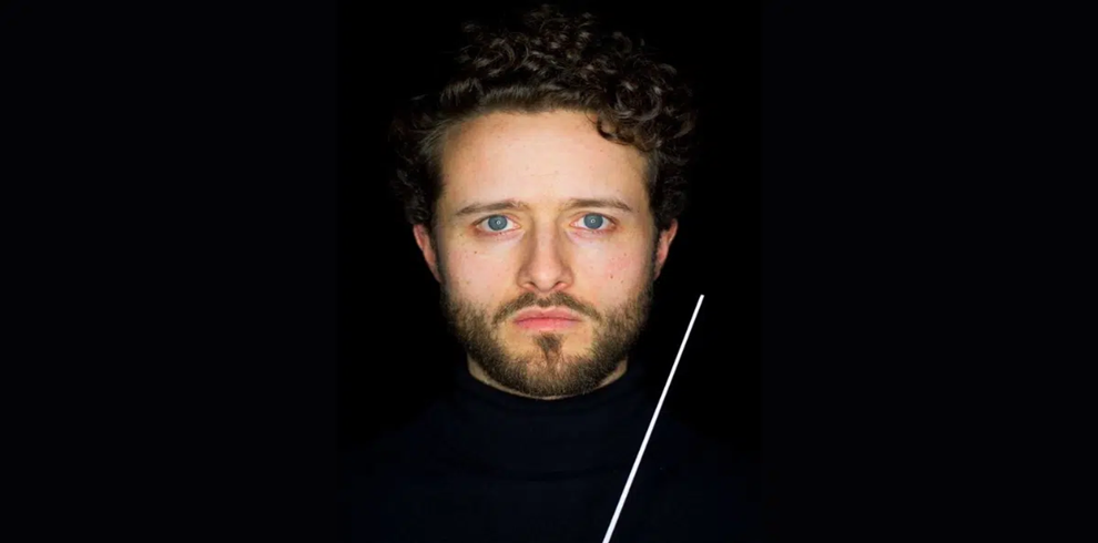 Studio portrait of a person with short curly hair and a beard, facing directly toward the camera against a black background. The person is wearing a dark high-neck top and is illuminated by focused lighting that highlights the face. A thin white conductor’s baton is visible diagonally across the lower right side of the image. The composition is centered and minimalist, with strong contrast between the subject and the dark background.