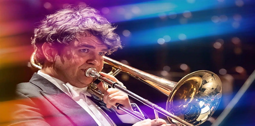 Close-up photograph of a musician playing a brass trombone during a live performance. The musician is wearing formal concert attire, including a dark jacket and white shirt. The trombone is prominently featured in the foreground, with the instrument’s polished bell reflecting stage lights. The background is softly blurred with blue, purple, and gold lighting.