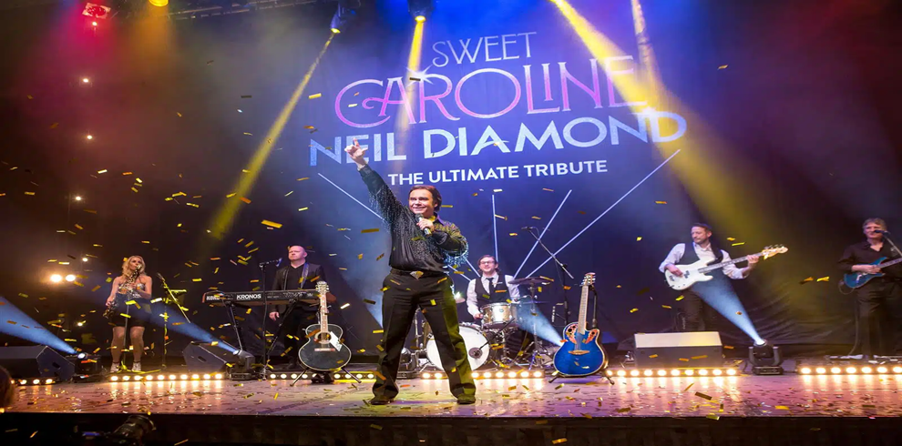 Promotional stage performance image for “Sweet Caroline: Neil Diamond – The Ultimate Tribute.” A live band performs on a brightly lit stage filled with colorful spotlights in blue, gold, red, and purple tones. At center stage, a performer in dark clothing stands with one arm raised while holding a microphone. Gold confetti falls across the stage and audience area. Surrounding musicians play keyboards, drums, bass guitar, and electric guitar, with several instruments visible on stands. Large text projected on the backdrop reads, “Sweet Caroline Neil Diamond The Ultimate Tribute.” Bright stage lighting, illuminated floor lights, and scattered confetti create a celebratory concert atmosphere.