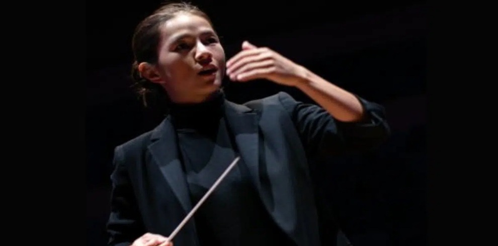 A person stands on a dimly lit stage holding a conductor's baton and gesturing with one raised hand. The person is dressed in a dark blazer and black top, illuminated by a spotlight against a dark background, suggesting an orchestral or musical performance setting.