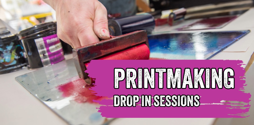 Close-up photograph of a hand using a printmaking roller covered in bright pink ink on a work surface. Printmaking tools and ink containers are visible in the blurred background. Overlaid on a purple paint-stroke graphic is the text: “PRINTMAKING” and “DROP IN SESSIONS” in large white lettering. The image promotes family-friendly printmaking activities.