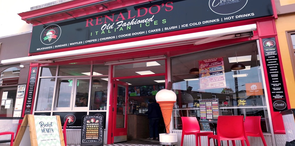 Front view of Renaldo’s Old Fashioned Italian Ices shop, featuring a red-and-black storefront with large windows and outdoor seating. A large sign above the entrance advertises Italian ices, milkshakes, sundaes, waffles, crepes, churros, cookies, cakes, slush drinks, and hot drinks. A decorative oversized ice cream cone stands outside the entrance beside several red chairs and tables. Promotional signs and menu boards are visible through the windows, creating a bright and welcoming café atmosphere.