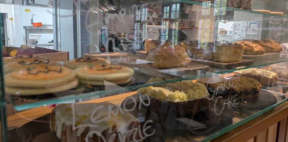Glass display case inside a café showing a selection of cakes, pastries, and baked goods. Reflections from the café interior are visible on the glass, with serving counters and seating in the background.