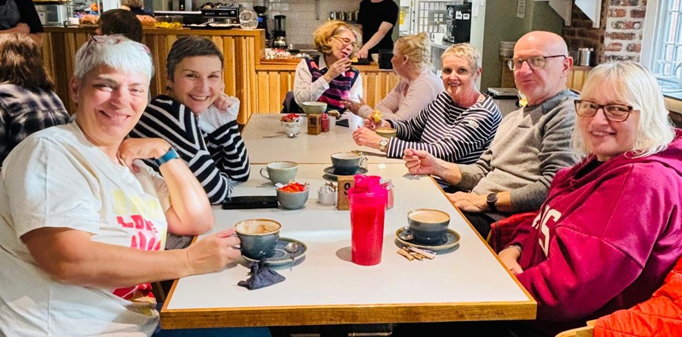 Several people gathered around a table in a café, enjoying drinks and conversation. The table is set with coffee cups, plates, and a bright red water bottle, creating a welcoming community setting.