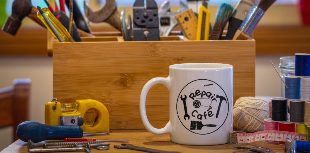 Promotional image for a Repair Café event. A wooden toolbox filled with repair and DIY tools, including hammers, spanners, pliers, rulers, paintbrushes and screwdrivers, sits on a table in the background. In the foreground is a white mug printed with the “Repair Café” logo, beside balls of twine, sewing thread spools, a measuring tape, a spirit level and hand tools. The image features warm lighting and a workshop setting, highlighting repair, mending and reusing items. The words “One Small Thing” appear in white text in the upper left corner.