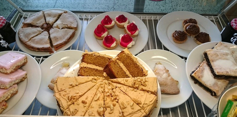 A display case filled with assorted baked treats on white plates. In the center foreground is a large round cake sliced into wedges and topped with a light caramel-colored frosting and crushed nuts. Behind it are slices of layered spice cake and iced pastries. Other plates hold a powdered sugar-topped pie or cake, pink iced traybakes, small tartlets with red jelly and cream, chocolate-topped mini pastries, and rectangular fruit-filled bars dusted with icing sugar. The desserts are arranged neatly on metal shelving inside a bakery or café display cabinet.
