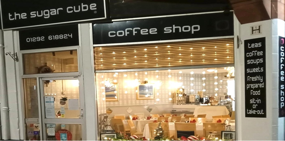 The Sugar Cube 1 Front view of The Sugar Cube coffee shop at night, with a black-and-white storefront and brightly lit interior visible through a large window. The signage reads “the sugar cube” and “coffee shop,” with a phone number displayed above the entrance. Inside, tables are set with white tablecloths and red napkins, and a service counter can be seen at the back. Decorative string lights hang across the front window, creating a warm glow. A vertical sign beside the window advertises “Teas, coffee, soups, sweets, freshly prepared food, sit-in or take-out.” The exterior is clean and welcoming, with festive decorations visible along the bottom of the window display.