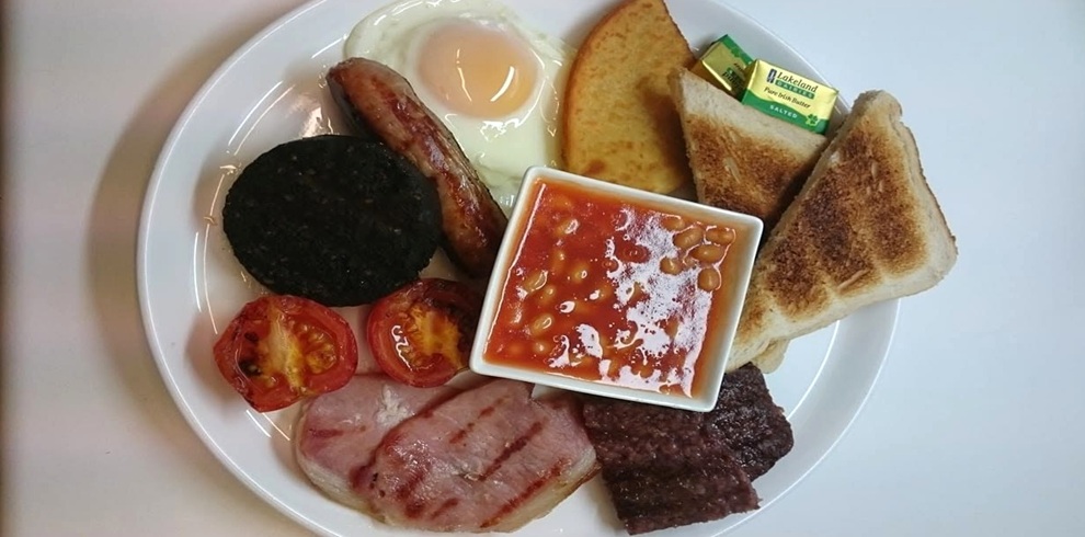 A full cooked breakfast served on a white plate against a plain white background. The plate includes a fried egg, a grilled sausage, a slice of black pudding, grilled bacon, a square sausage patty, toasted bread cut into triangles, a slice of buttered toast, and grilled tomato halves. In the center sits a small square dish filled with baked beans in tomato sauce. Individual butter portions rest beside the toast. The meal is neatly arranged and photographed from above, highlighting the variety of traditional cooked breakfast items.