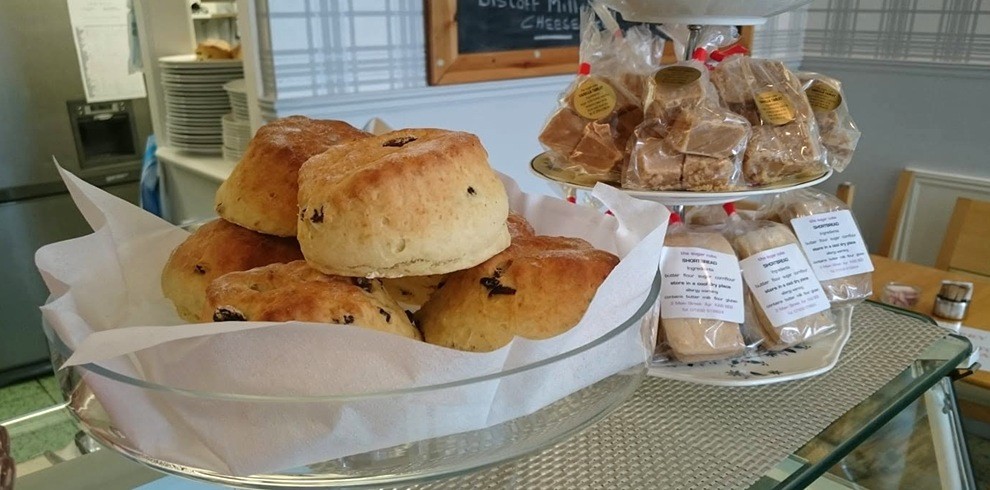 A café display counter featuring a large glass bowl lined with white paper and filled with freshly baked fruit scones. Beside it, a two-tier stand holds individually wrapped baked goods and shortbread-style treats in clear packaging with product labels attached. The display sits on a glass countertop in a bright café setting. In the background are stacked white plates, light-colored patterned wallpaper, dining chairs and tables, and a chalkboard sign with partially visible text. The baked items are arranged neatly, creating an inviting homemade bakery display.
