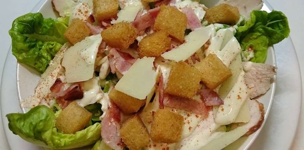 Close-up view of a Caesar-style salad served in a white bowl. The salad is made with crisp green lettuce leaves, sliced grilled chicken, strips of bacon, golden croutons, and shaved Parmesan cheese. A creamy dressing is drizzled generously over the ingredients and lightly sprinkled with seasoning. The fresh green lettuce frames the edges of the bowl, while the croutons and cheese are distributed across the top, creating a colourful and textured presentation against a plain white background.