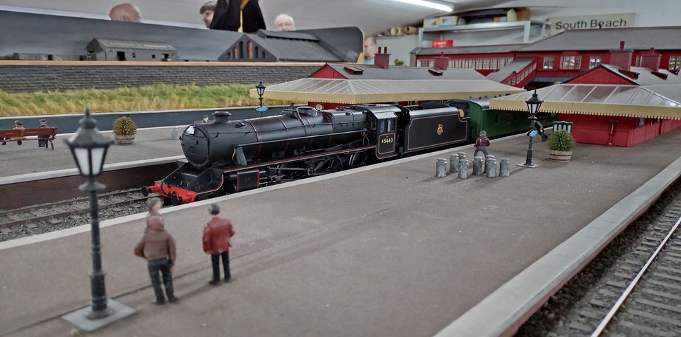 A detailed model railway station display featuring a black steam locomotive numbered 45443 stopped at a platform beside red station buildings. Miniature figures stand on the platform near lamp posts and luggage trolleys, while multiple railway tracks run through the foreground. In the background, additional model structures and a station sign reading “South Beach” are visible inside an exhibition setting.