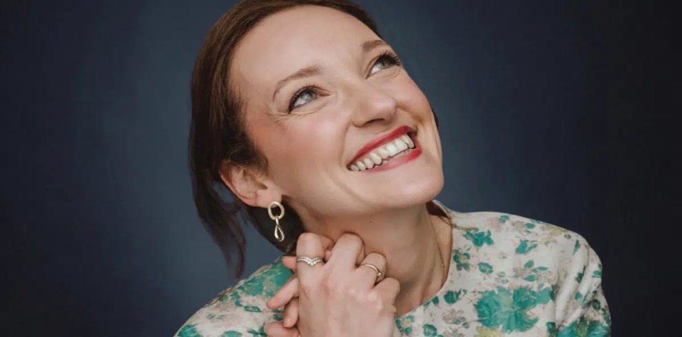 Head-and-shoulders portrait of a person with dark hair against a deep blue background. The person is wearing a light-coloured top with a green and turquoise floral pattern and dangling earrings. Hands are gently clasped beneath the chin, with several rings visible on the fingers. The face is turned upward and slightly to one side, showing a broad smile. Soft, even lighting highlights the facial features and patterned clothing, while the background remains smooth and uncluttered.
