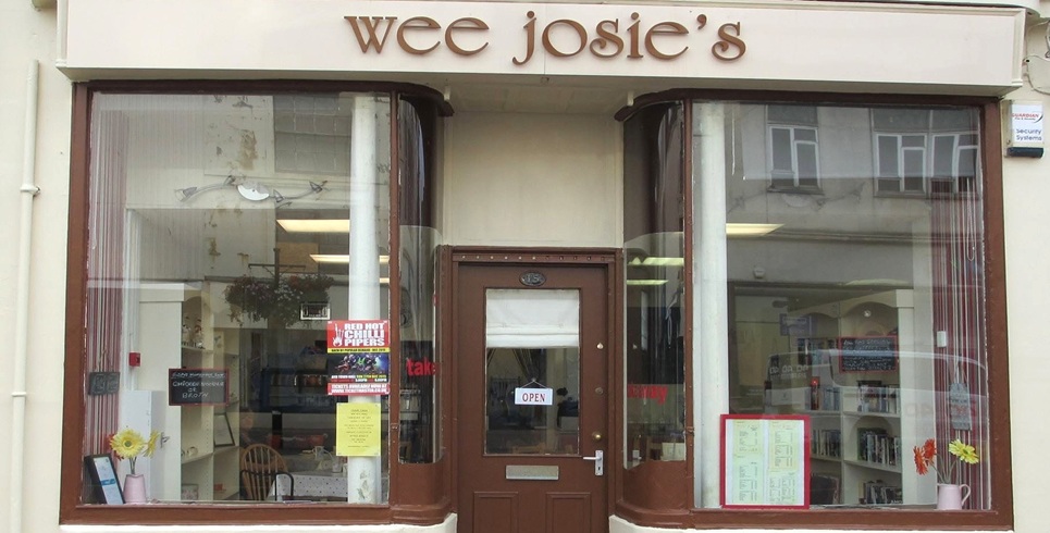 Front view of a small storefront named “wee josie’s” with large display windows on either side of a central brown entrance door. The door has an "OPEN" sign, and the windows contain books, posters, notices, and floral decorations. A menu board is displayed in the right window, while additional signs and a chair are visible in the left window. The cream-colored shopfront features curved window frames and reflects nearby buildings in the glass.
