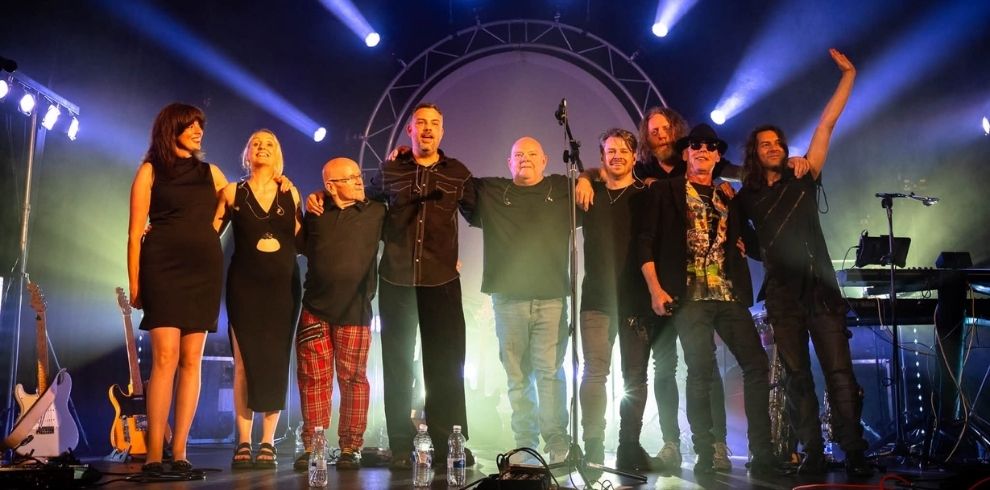 A group of nine performers stand across a concert stage with arms around each other beneath bright blue and white stage lights. Musical instruments, including guitars and keyboards, are positioned at the sides of the stage. Several water bottles and microphone stands are visible at the front, with a large circular backdrop behind the performers.