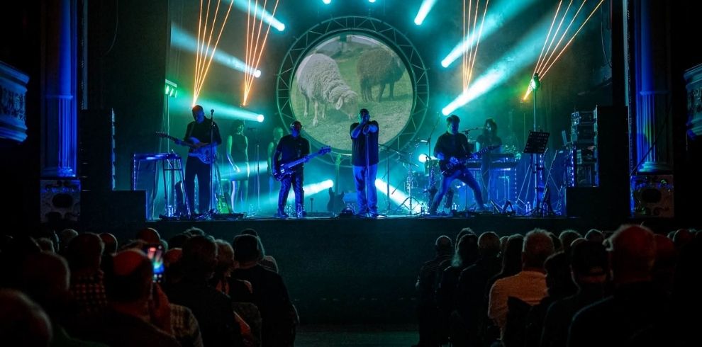 A live band performs on a theatre stage illuminated by blue, green, and white lighting effects. Several musicians are silhouetted against a large circular screen displaying an image of sheep. Audience members are seated in rows in the foreground facing the stage.