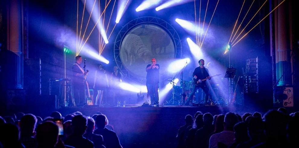 A live band performs on a theatre stage illuminated by blue, and white lighting effects. Several musicians are silhouetted against a large circular screen displaying an image of sheep. Audience members are seated in rows in the foreground facing the stage.