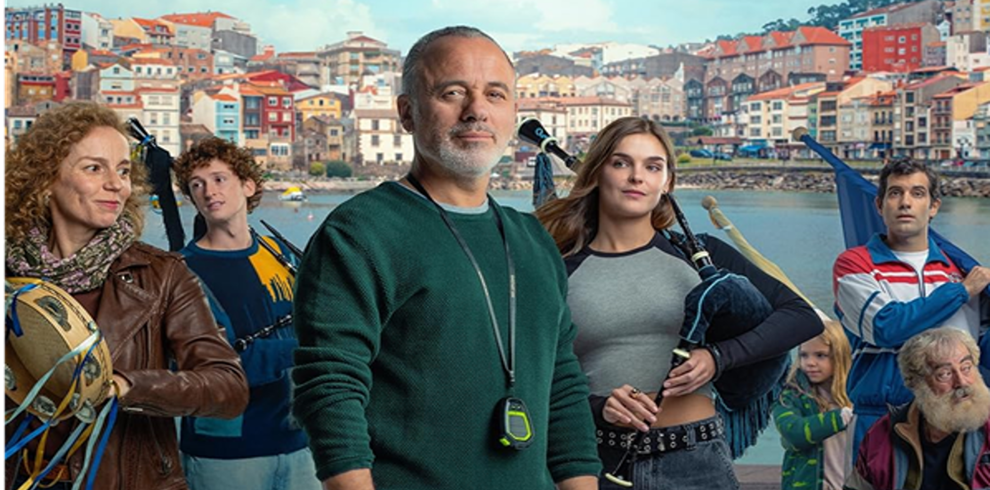 Promotional image for The Rondalla. A group of people stand along a waterfront with a densely built coastal town rising on the hillside behind them. In the centre foreground, a person wearing a dark green sweater stands facing the camera with a whistle or stopwatch hanging around their neck. Surrounding figures hold traditional musical instruments, including bagpipes and a tambourine, suggesting a community folk music ensemble. The harbour and shoreline are visible in the background, with colourful buildings featuring red-tiled roofs stretching across the hillside. The image combines maritime scenery with musical imagery, reflecting the film's themes of community, tradition and renewal.