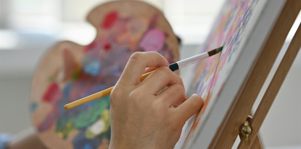 Close-up of a hand holding a paintbrush while painting a colourful canvas on an easel. A paint palette covered in bright mixed colours is blurred in the background, highlighting a creative art workshop or painting class.