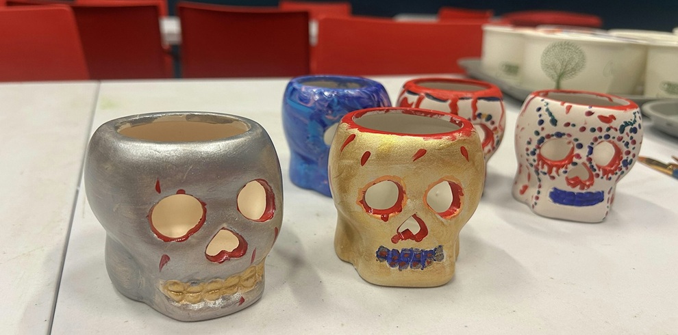 Several hand-painted ceramic skull lanterns in silver, gold, blue and white sit on a table during a pottery painting workshop. The decorative skulls feature colourful painted details around the eyes, nose and teeth, with red chairs visible in the background.