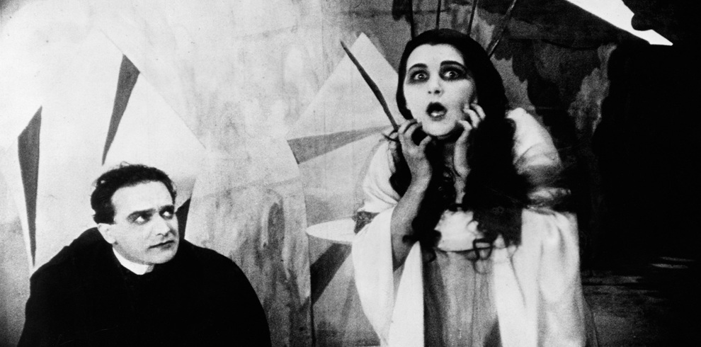 Black-and-white still from The Cabinet of Dr. Caligari. In the foreground, two figures stand against a backdrop of sharply angled, abstract set pieces characteristic of German Expressionist design. On the left, a figure dressed in dark clothing sits or crouches while looking toward the other figure. On the right, a figure in a light-coloured flowing gown stands with hands raised near the face. The background is filled with painted geometric shapes, distorted architectural forms and dramatic contrasts of light and shadow. The highly stylised composition creates an eerie, dreamlike atmosphere that reflects the film's themes of mystery, madness and illusion.