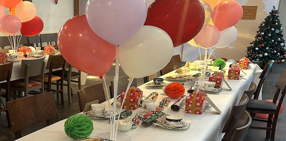 Long banquet-style table decorated for a festive holiday gathering, set with teacups, plates, crackers, and small wrapped gift boxes. Clusters of red, white, and pink balloons are arranged along the table, with colorful paper decorations placed between settings. Additional decorated tables are visible in the background, and a Christmas tree stands in the corner of the room. Warm indoor lighting creates a welcoming seasonal atmosphere.