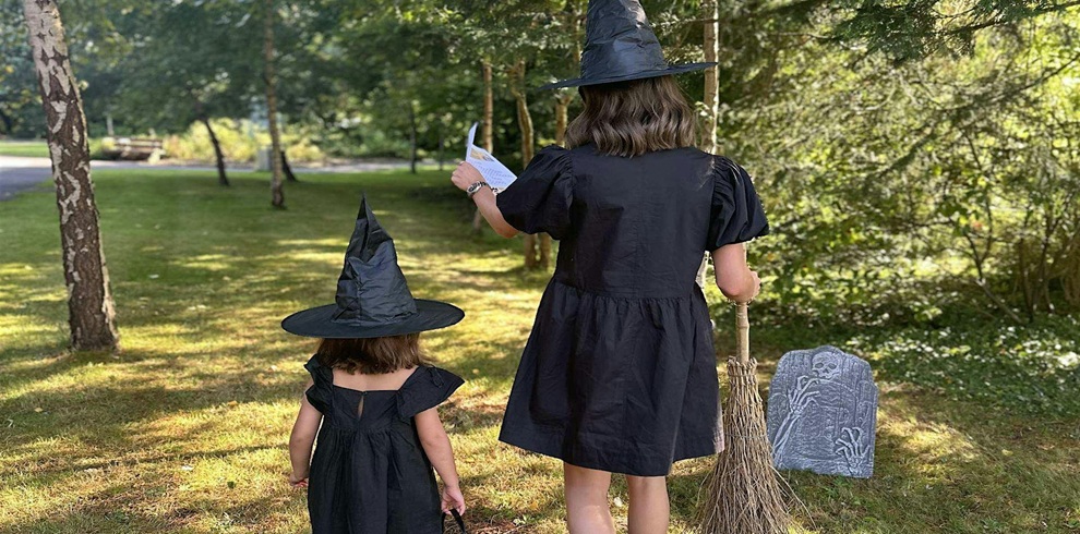 Two people in black witch costumes walk through a sunlit garden, following a trail activity. One carries a sheet of paper while the other stands nearby. A broomstick and a decorative gravestone prop add a spooky Halloween atmosphere among the trees and grassy grounds.