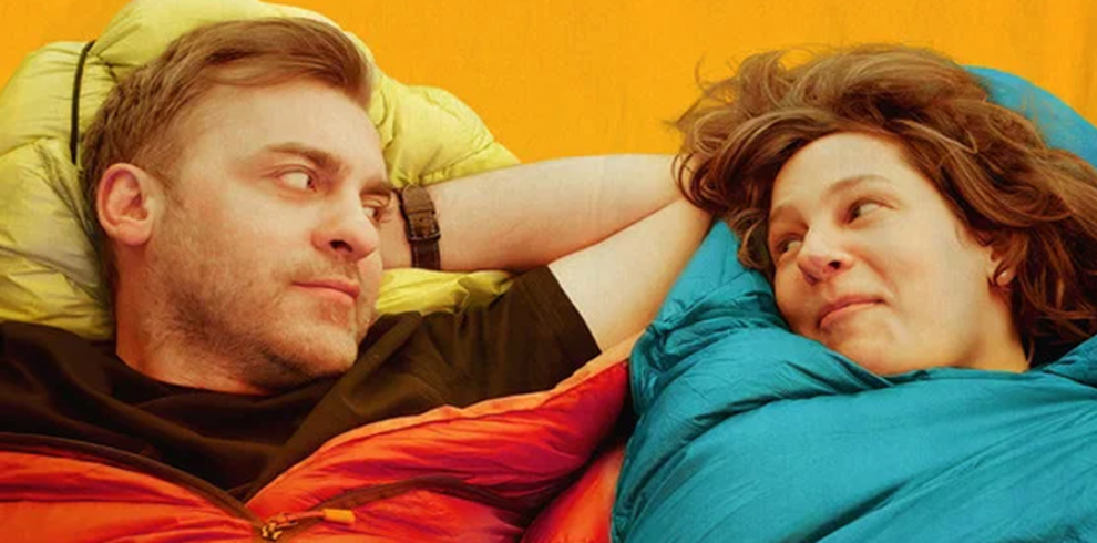 Promotional image for This Is Not My Film. Two people lie side by side facing one another against a bright yellow background. Each person is wrapped in a colourful sleeping bag, one in shades of red and orange and the other in bright blue, with their heads resting on camping pillows. The composition is closely cropped, focusing on the two figures and creating a sense of intimacy and conversation. The vivid contrasting colours and minimalist background give the image a warm, playful and contemporary appearance, suggesting themes of relationships, self-discovery and personal journeys.