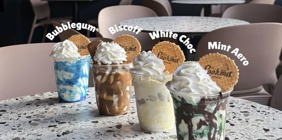 Four takeaway soft-serve ice cream desserts displayed on a speckled café table inside The Lookout Troon. The flavours, labelled above each cup, are Bubblegum, Biscoff, White Choc and Mint Aero. Each dessert is topped with whipped cream and a branded biscuit, with colourful sauces and toppings matching the flavour. Soft pink café chairs and tables are visible in the background.